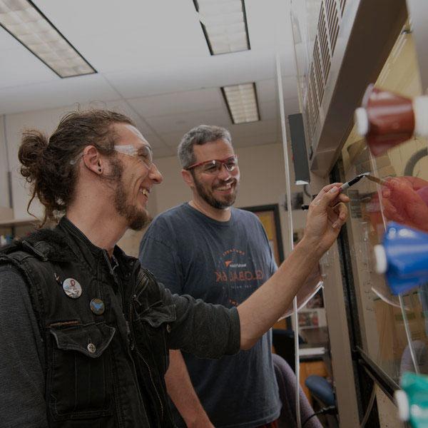 Chemistry student writes on the board while discussing with his professor. Both are wearing safety glasses.